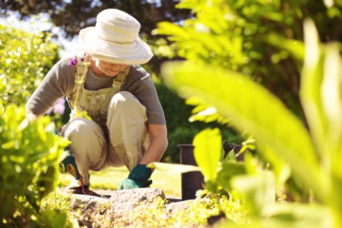 A woman wearing a wide-brimmed sun hat, glasses, a floral apron, and gardening gloves is kneeling on a well-maintained lawn in a residential garden, tending to flower beds with a trowel and small garden tools. The garden features a lush, green grassy area in the foreground, bordered by neatly edged flower beds containing various plants and shrubs. In the background, there are tall trees and a hedge that provides privacy and natural border to the outdoor space. The sunlight creates bright highlights and soft shadows, indicating a clear, sunny day. The scene reflects regular outdoor maintenance, with the garden clean and organized, aligning with professional gardening services in Sutton that focus on landscape care, planting, and outdoor upkeep, ensuring healthy plants and visually appealing garden environments.