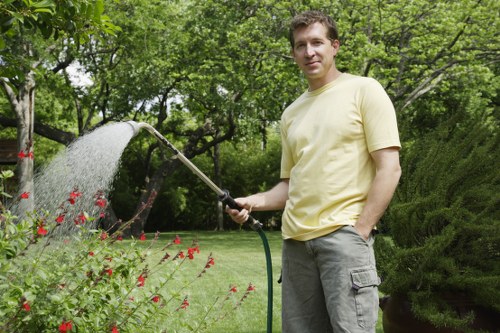 A man standing in a well-maintained garden, holding a garden hose sprayer and watering plants. The garden features a lush, green lawn bordered by flower beds with red blooms, dense trees and shrubs in the background. The scene is outdoors with natural daylight, and the overall setting appears tidy and cultivated, indicative of professional gardening and lawn care services typically offered in Sutton. The garden landscape includes a variety of greenery, with trimmed bushes and a spacious, landscaped yard suitable for outdoor maintenance and gardening projects conducted by Gardening Services Sutton.