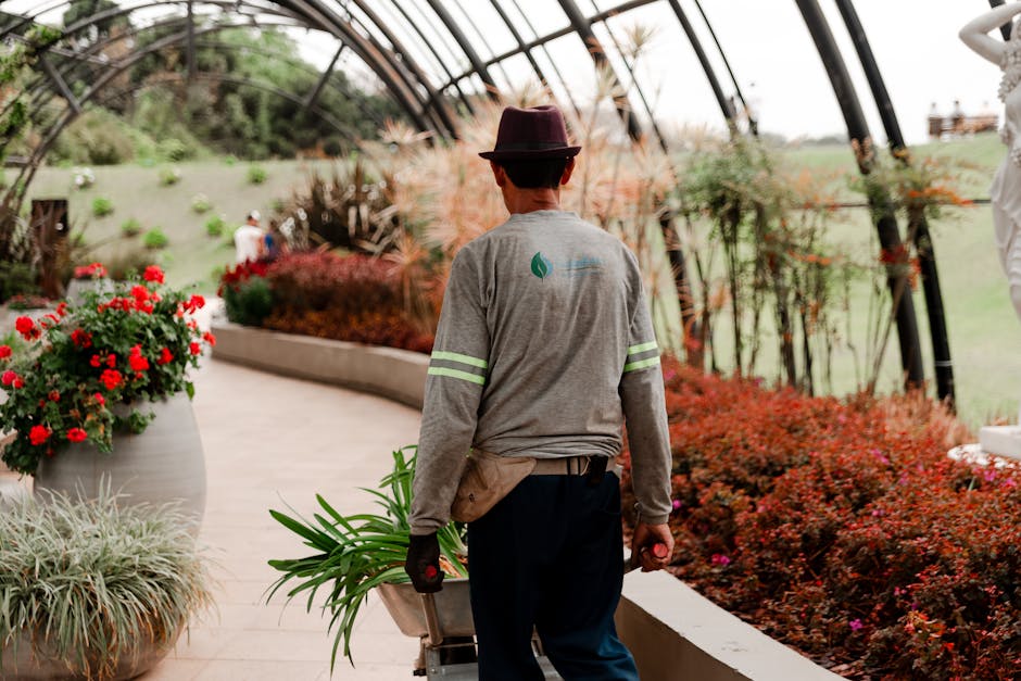 A gardener wearing a grey long-sleeve shirt with reflective stripes on the sleeves, dark pants, and a brown hat is pushing a wheelbarrow through a spacious, modern greenhouse. The greenhouse features 