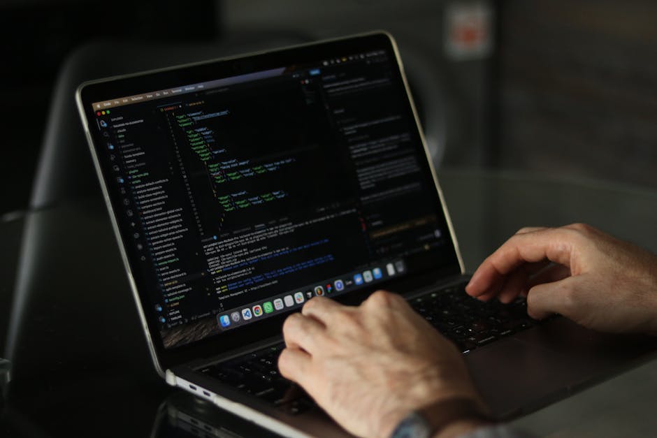 A person working on a laptop with a dark-themed code editor open, displaying lines of colorful programming code, placed on a reflective black surface. The individual's hands are visible, with the righ