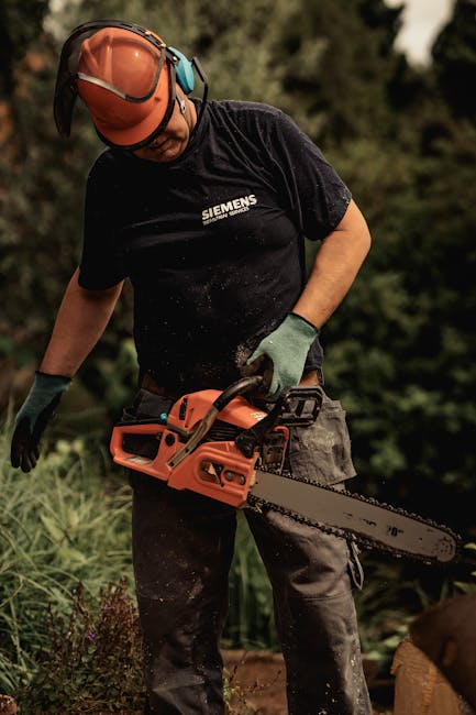 A male gardener wearing a safety helmet and protective gloves is operating an orange chainsaw on a garden lawn. He is dressed in a black t-shirt with the Siemens logo, and grey work trousers. The surr